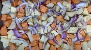 Chopped sweet potatoes, red onions, garlic, and herbs seasoned for roasting in a baking tray.